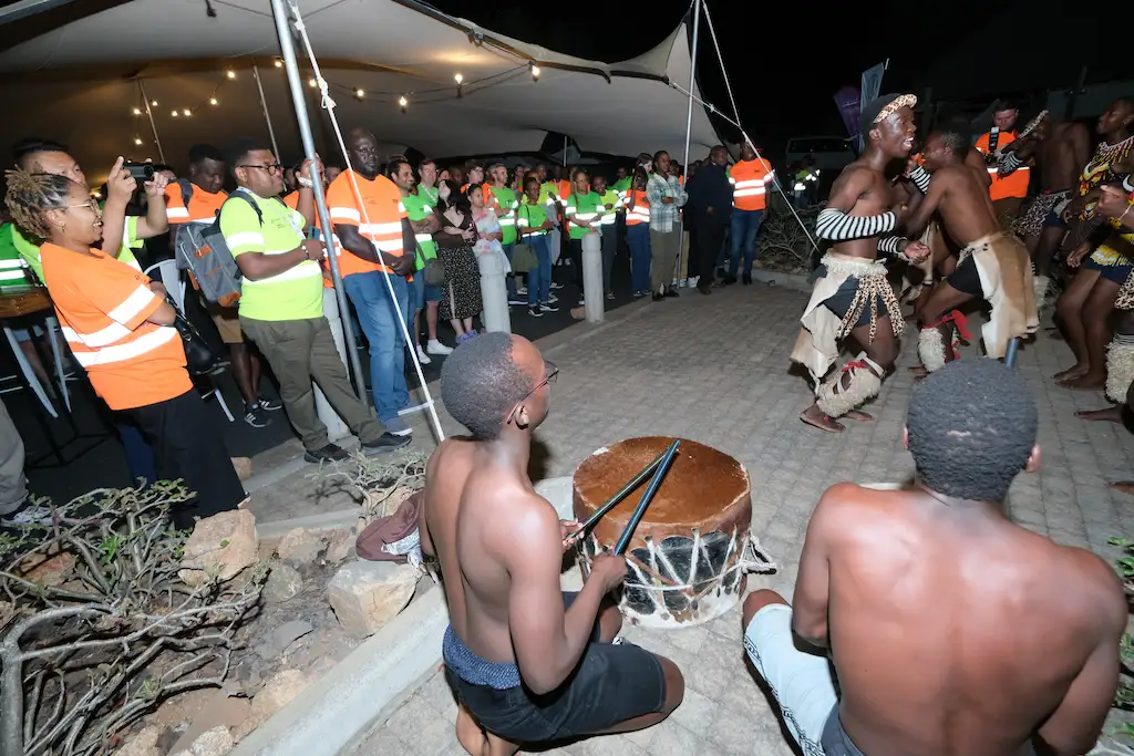 Traditional African dancers performing with drummers while a diverse audience in safety vests watches at an outdoor event.