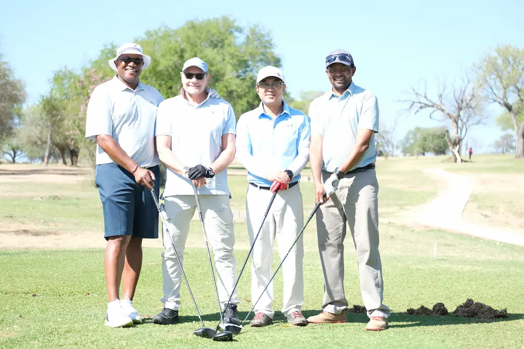 Four men posing with golf clubs on a golf course during a sunny day, dressed in casual golf attire.