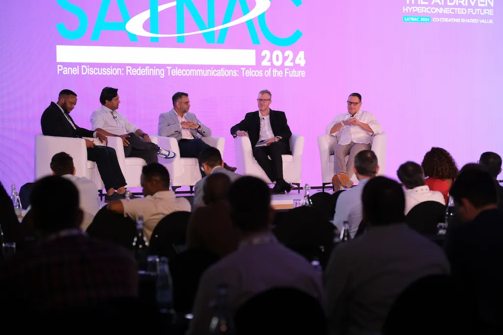 Five male panelists in armchairs on a brightly lit stage during a panel discussion at the SATNAC 2024 conference. The screen behind them reads, "Panel Discussion: Redefining Telecommunications: Telcos of the Future.