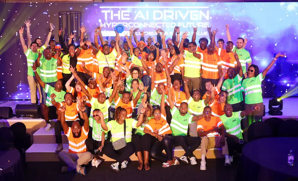 A large, diverse group of enthusiastic conference attendees and staff in bright orange and green t-shirts, posing for a group photo on a stage. The screen behind them reads, "THE AI DRIVEN HYPERCONNECTED FUTURE.
